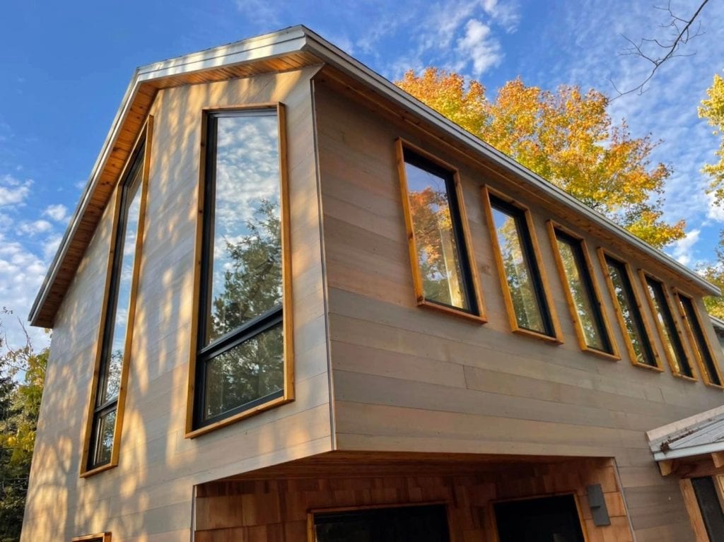 Modern wooden house exterior featuring large windows and a contemporary design, surrounded by autumn trees under a blue sky, showcasing professional painting and decorating services.